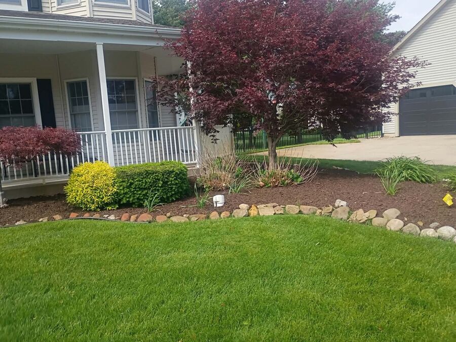 Wide view of a curved mulched bed with river-rock edging, layered shrubs, and ornamental grasses beside a lawn.