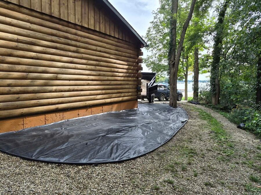Black landscape fabric spread over graded soil beside a lakeside log building, shaped around mature trees.