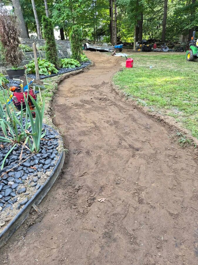 Graded soil corridor curving between a planted slope and lawn, with wheelbarrows and equipment in the background.